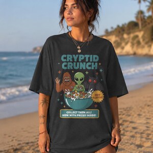 May include: A black t-shirt featuring a graphic of a brown Bigfoot, a green alien, and a black bat-like creature eating a bowl of colourful cereal. The text reads CRYPTID CRUNCH, NEW FLAVOR, COLLECT THEM ALL! NOW WITH PRIZES INSIDE! with small stars.