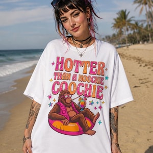 Pode incluir: Uma t-shirt branca de manga curta com um gráfico de um gorila castanho a usar óculos de sol e um colete cor-de-rosa, sentado numa boia cor-de-rosa e laranja enquanto segura uma bebida tropical. O texto diz HOTTER THAN A HOOCHIE COOCHIE.