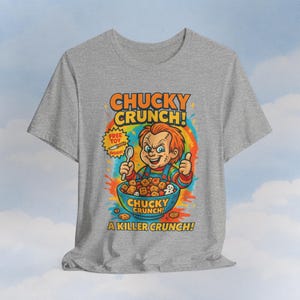 Könnte beinhalten: Graues T-Shirt mit einem farbenfrohen Grafik von Chucky, der einen Löffel über einer Müslischüssel hält. Der Text lautet "CHUCKY CRUNCH!" und "A KILLER CRUNCH!" mit einer "FREE TOY INSIDE!" Grafik.
