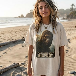 May include: A beige short-sleeved t-shirt featuring a rectangular graphic print of a brown, furry Bigfoot creature in a forest setting. The text below the creature reads PACIFIC NORTHWEST BIGFOOT in bold, dark, capitalized lettering.
