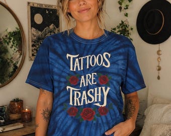 May include: A blue tie-dye t-shirt featuring the white text "TATTOOS ARE TRASHY" printed in the center. The design includes a decorative arrangement of red roses with green leaves positioned above and below the text on the front of the shirt.