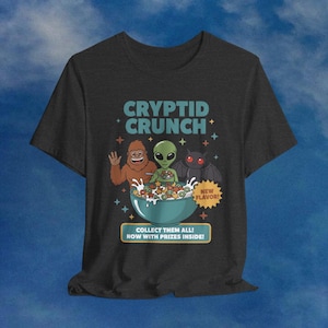 Könnte beinhalten: Dunkelgraues T-Shirt mit einem Cartoon-Design einer Müslischüssel mit einem Bigfoot, einem Alien und einer Fledermaus. Der Text "CRYPTID CRUNCH" steht über der Schüssel, darunter "NEW FLAVOR!" und "COLLECT THEM ALL! NOW WITH PRIZES INSIDE!"