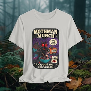 Könnte beinhalten: Weißes T-Shirt mit einem Aufdruck eines schwarzen Mothman-Charakters mit roten Augen und Flügeln, der Müsli isst. Das Design enthält den Text "Mothman Munch" und "A Cryptid You Can Crunch!" mit einem kostenlosen, im Dunkeln leuchtenden Augenaufkleber.