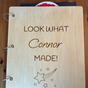May include: A light-colored wooden scrapbook with metal ring binding. The cover is engraved with the words "LOOK WHAT Connor MADE!" and a shooting star design with star accents. The scrapbook is open.