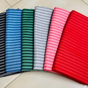 May include: Seven folded fabric pieces in various colors: black, blue, green, gray, pink, and red. Each piece features horizontal stripes. The fabrics are neatly arranged, showcasing a range of hues and patterns.