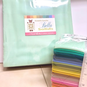 May include: A stack of colorful fabric squares, with a ruler for measurement, next to a large, mint green fabric square. The label reads "Elizabeth's Favorites Bella Solids" and "moda FABRICS SUPPLIES".