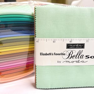 May include: Stacks of colorful fabric squares, including shades of green, blue, yellow, pink, and orange. A stack of light green fabric squares is in focus, with a white label that reads "Elizabeth's Favorites Bella Solid" by Moda.