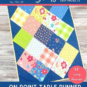 May include: A colorful table runner with a patchwork design. The runner features various patterned squares in shades of blue, yellow, pink, and green, with floral and gingham patterns. The text "ON POINT TABLE RUNNER" is displayed. The runner is 53 inches long.