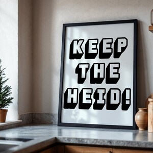 以下が含まれることがあります： 白い背景に、太字の黒い3Dスタイルの文字で「KEEP THE HEID！」と書かれた額入りのプリント。 黒いフレームは長方形です。 プリントはキッチンのカウンタートップに表示されています。