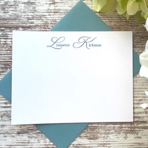 May include: A white stationery card with the name "Lynnette Kirkman" printed in a delicate blue script, resting on a teal envelope. The background features a rustic wooden surface and white flowers.