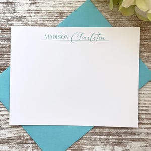 May include: A white stationery card with the name "MADISON Charlton" printed in a teal script font, resting on a teal envelope. The stationery is set against a wooden background, with white and green flowers in the upper right corner.