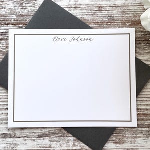 May include: A white stationery card with a thin gray border and the name "Dave Johnson" in script. The card rests on a dark gray envelope, with a white flower in the background. The stationery is on a white wooden surface.