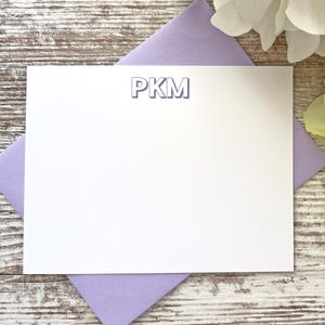 May include: A white stationery card with the initials "PKM" in a light blue font, resting on a lavender envelope. The background features a rustic, white-washed wooden surface, with white and green flowers in the upper right corner. This stationery is ideal for personalized notes.