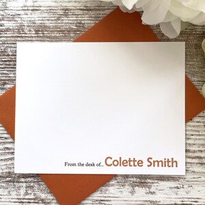 May include: A white stationery card with the text "From the desk of... Colette Smith" in brown font, resting on a rust-colored envelope. The background is a weathered, light-colored wooden surface, with white and green floral accents.