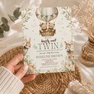 May include: A baby shower invitation featuring a hot air balloon with two teddy bears, a bunny, and a deer. The invitation has the text "We can bearly wait TWIN" and details for the event.