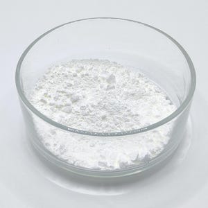May include: A clear glass bowl containing a white, powdery substance. The bowl is circular and rests on a white surface. The substance looks finely ground and has a slightly textured appearance.