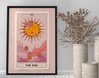 Peut inclure: Impression encadrée d'une carte de tarot représentant le soleil souriant avec des détails floraux. La carte est sur un fond rose avec des nuages et les mots "THE SUN". Le cadre est noir.