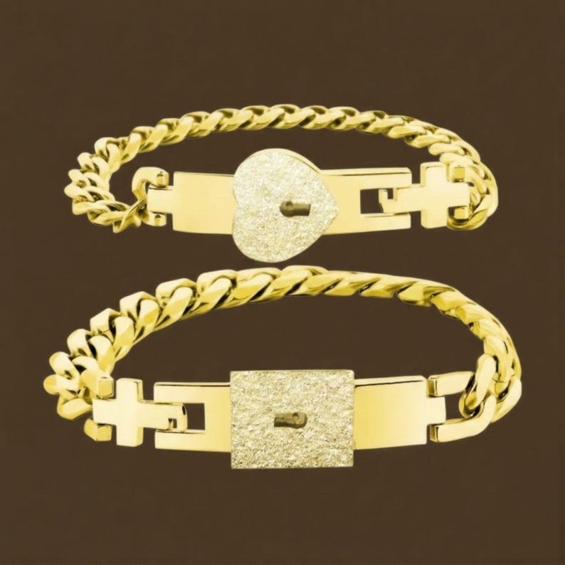 Puede incluir: Dos pulseras doradas con dise&ntilde;o de eslabones de cadena. Una pulsera presenta un candado en forma de coraz&oacute;n, y la otra tiene un candado cuadrado. Ambos candados tienen una apariencia texturizada y brillante. Las pulseras se muestran sobre un fondo marr&oacute;n.