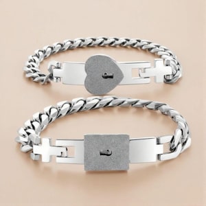 Peut inclure: Deux bracelets argentés avec un motif de chaîne. L'un présente un cadenas en forme de cœur, l'autre un cadenas carré. Les deux cadenas ont une serrure. Les bracelets sont présentés sur une surface beige clair.