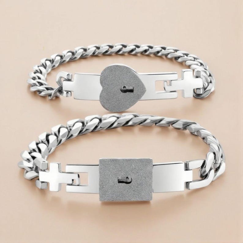 Puede incluir: Dos pulseras plateadas con dise&ntilde;o de cadena. Una pulsera presenta un candado en forma de coraz&oacute;n y la otra un candado cuadrado. Ambos candados tienen un dise&ntilde;o de ojo de cerradura. Las pulseras se exhiben sobre una superficie beige claro.