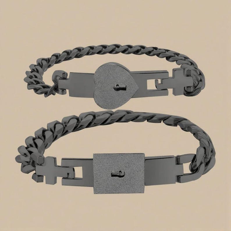 Puede incluir: Dos pulseras negras a juego. Una presenta un candado en forma de coraz&oacute;n, la otra un candado cuadrado. Ambas tienen un dise&ntilde;o de eslabones de cadena y un detalle de ojo de cerradura. Las pulseras se muestran sobre un fondo neutro.