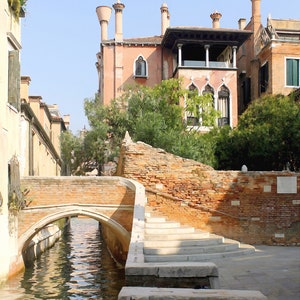 May include: A brick bridge over a canal in Venice, Italy. The bridge has a stone archway and steps leading up to it. The canal is narrow and the water is clear. The building in the background is a pink building with a balcony and a tiled roof. The building has a sign that says "GRANAI".