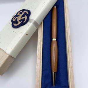 May include: A wooden pen with gold-coloured tip and accents, presented in a wooden box lined with blue fabric. The box is partially open, revealing the pen. The lid is decorated with a blue and gold knot design.