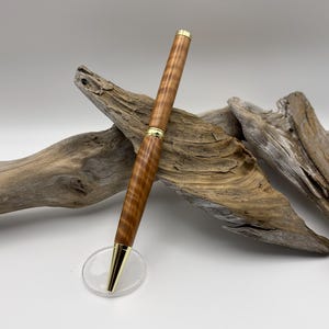 May include: A handcrafted wooden pen with a gold-colored tip and accents. The pen features a light brown, patterned wood grain and rests on a clear acrylic stand. The pen is approximately 6 inches long.