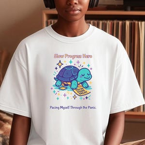 May include: A white short-sleeved t-shirt featuring a graphic of a blue turtle with a purple shell, surrounded by colorful stars. The text reads Slow Progress Here and Pacing Myself Through the Panic, with a small sign saying You're Doing Great.