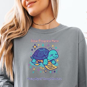 May include: A grey long-sleeve crewneck sweatshirt featuring a graphic of a teal turtle with a dark blue shell. The text reads Slow Progress Hero and Pacing Myself Through the Panic, with a small yellow sign saying You're Doing Great.