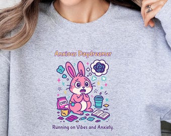 May include: A light grey crewneck sweatshirt featuring a graphic of a pink rabbit with the text Anxious Daydreamer and Running on Vibes and Anxiety. The illustration includes a bag of chips, a cup, a book, and various small colourful shapes.