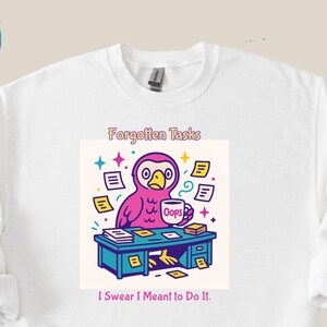 Puede incluir: Sudadera blanca con un diseño colorido. Muestra un pájaro rosa en un escritorio, rodeado de papeles, con el texto "Forgotten Tasks" y "I Swear I Meant to Do It." El pájaro sostiene una taza de café. El diseño incluye estrellas coloridas.