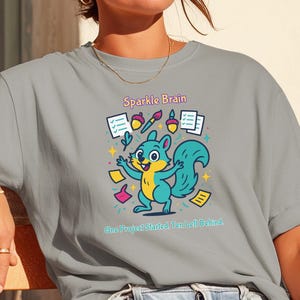 May include: A grey t-shirt featuring a colorful graphic of a teal squirrel surrounded by floating items including checklists, acorns, and a paintbrush. The text reads Sparkle Brain above the squirrel and One Project Started Ten Left Behind below.