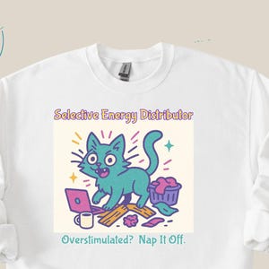 Puede incluir: Sudadera blanca con un gráfico colorido de un gato turquesa con el texto "Selective Energy Distributor" y "Overstimulated? Nap It Off." El gato está representado con un portátil, una taza de café y una cesta de lavandería.