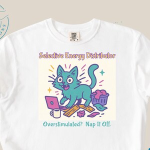 Puede incluir: Camiseta blanca con un diseño de gato de dibujos animados. El texto dice "Selective Energy Distributor" y "Overstimulated? Nap It Off." El gato turquesa está representado con un portátil, una taza de café y una cesta de la ropa, todo en un diseño colorido.
