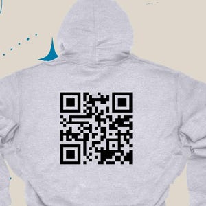Op de afbeelding: Een lichtgrijze hoodie met een capuchon en een grote zwarte QR-code op de achterkant. De hoodie heeft een eenvoudig ontwerp en is gemaakt van een zacht materiaal. Het Anillav Creations-logo staat linksboven.