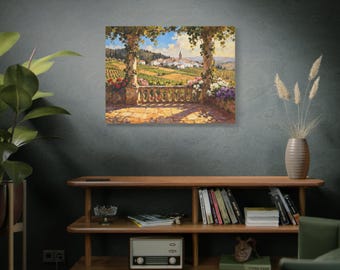 May include: A framed canvas print of a scenic landscape. The artwork depicts a sunny vista of a European village, vineyards, and a stone balcony with flowers. The painting is displayed on a dark gray wall above a wooden shelf.