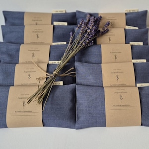 Könnte beinhalten: Anordnung von indigofarbenen Augenpolstern, jeweils mit einem braunen Papierband umwickelt. Ein Bündel getrockneter Lavendel ist mit Bindfaden gebunden und liegt obenauf. Die Papierbänder haben Text und ein Logo.
