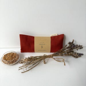 Könnte beinhalten: Ein rotes Augenkissen mit einem braunen Papieretikett, einer kleinen Glasschale mit Getreide und einem Bündel getrockneten Lavendels. Auf dem Etikett steht "Augenkissen" und "LISA YOGA". Das Augenkissen ist rechteckig.