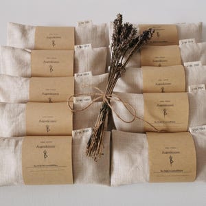 Op de afbeelding: Een verzameling beige linnen oogkussens, elk verpakt met een bruine papieren band met tekst en een logo. Een bosje gedroogde lavendel, vastgebonden met touw, ligt bovenop de kussens. De kussens zijn in nette rijen gerangschikt.