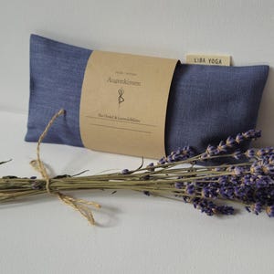 Op de afbeelding: Een blauw oogkussen met een bruin papieren label en een bosje gedroogde lavendel. Op het label staat "Augenkissen" en "LIBA YOGA". De lavendel heeft paarse bloemen en is vastgebonden met touw.