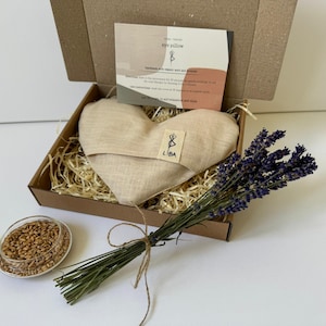 Könnte beinhalten: Ein herzförmiges, beiges Augenkissen liegt in einer Pappschachtel mit Stroh. Ein Lavendelstrauß und eine kleine Schale mit Getreide befinden sich neben der Schachtel. Die Schachtel enthält auch eine Karte mit dem Text "eye pillow" und dem Markennamen "LIBA".