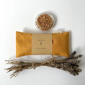 Könnte beinhalten: Senffarbenes Augenkissen mit braunem Papieretikett, einem kleinen Glasbehälter mit Körnern und einem Bündel getrockneten Lavendels. Das Etikett lautet "Augenkissen" und "Bio-Dinkel & Lavendelblüten". Ein kleines Etikett lautet "LIBA YOGA".