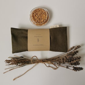 Może przedstawiać: Oliwkowa poduszka na oczy z brązową papierową etykietą, małą szklaną miseczką wypełnioną ziarnami i wiązką suszonej lawendy. Na poduszce na oczy widnieje napis "Augenkissen" i "Bio Dinkel & Lavendelblüten". Mała etykieta głosi "LISA YOGA".