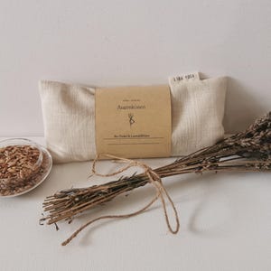 Może przedstawiać: Beżowa poduszka na oczy z brązową etykietą z napisem "Augenkissen" i pęczkiem suszonej lawendy. Mała szklana miseczka zawiera nasiona. Poduszka jest oznaczona "LISA YOGA".