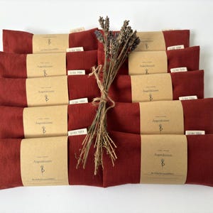 Könnte beinhalten: Eine Anordnung roter Augenpolster mit braunen Papieretiketten und einem Bündel getrockneten Lavendels. Die rechteckigen Augenpolster sind mit Papierbändern umwickelt. Die Etiketten tragen Text und ein Logo.