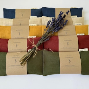 Könnte beinhalten: Eine Auswahl rechteckiger Augenpolster in verschiedenen Farben, darunter Marineblau, Creme, Gelb, Burgunderrot und Olivgrün. Jedes Kissen ist mit einem braunen Papierband und einem kleinen Etikett umwickelt. Ein Bündel getrockneter Lavendel ist mit Bindfaden gebunden.