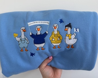 Può includere: Una felpa azzurra con un ricamo raffigurante quattro oche bianche. Un'oca indossa una salopette blu, una tiene uno striscione con la scritta Down Syndrome Awareness, una indossa una felpa gialla con cappuccio e una indossa berretto e sciarpa.
