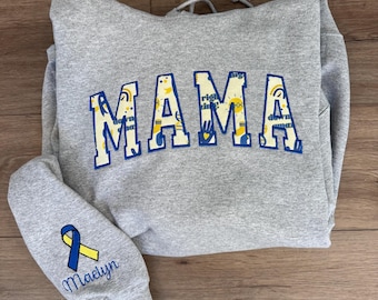 May include: A gray sweatshirt with the word "MAMA" in blue and yellow block letters. The sleeve features a blue and yellow ribbon and the name "Maelyn" in script. The sweatshirt is made of a soft material.