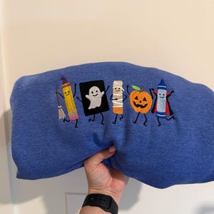 May include: A blue sweatshirt featuring an embroidered design of five cartoon characters: a pencil dressed as a witch, a ghost, a mummy, a jack-o'-lantern, and a blue crayon dressed as a vampire, all arranged in a horizontal line.